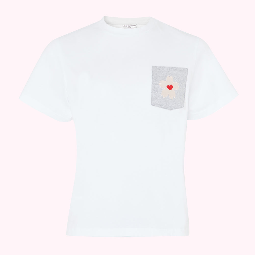 White t-shirt with grey pocket featuring a white flower applique and a red heart detail, on a pink background.