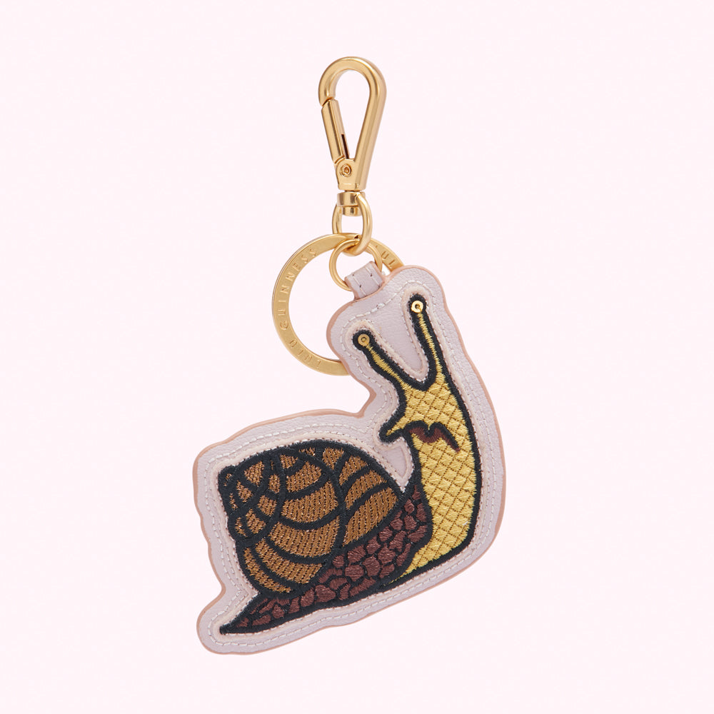 A pale pink leather keyring in the shape of a snail, with gold and brown embroidery detailing.