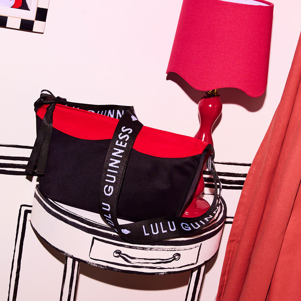 Black and red nylon crossbody bag with black and white branded 'Lulu Guinness' strap. Sitting on a black and white side table and next to a red lamp.