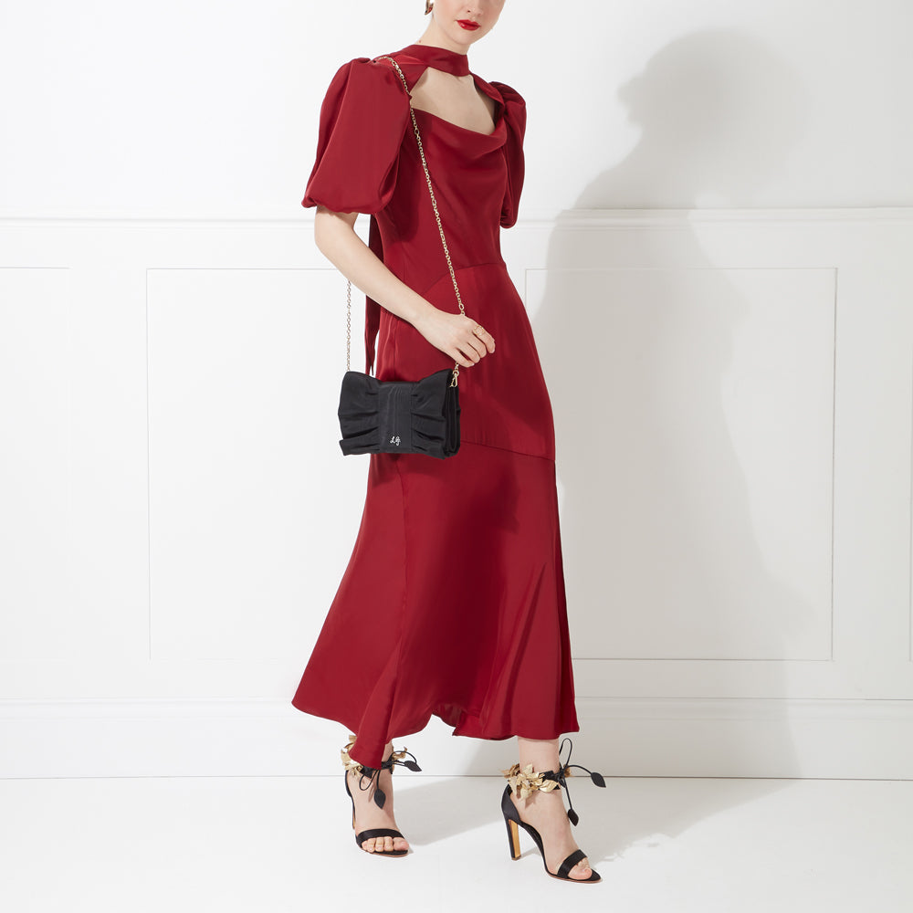 A woman in a burgundy maxi dress carries a black bow-shaped clutch.
