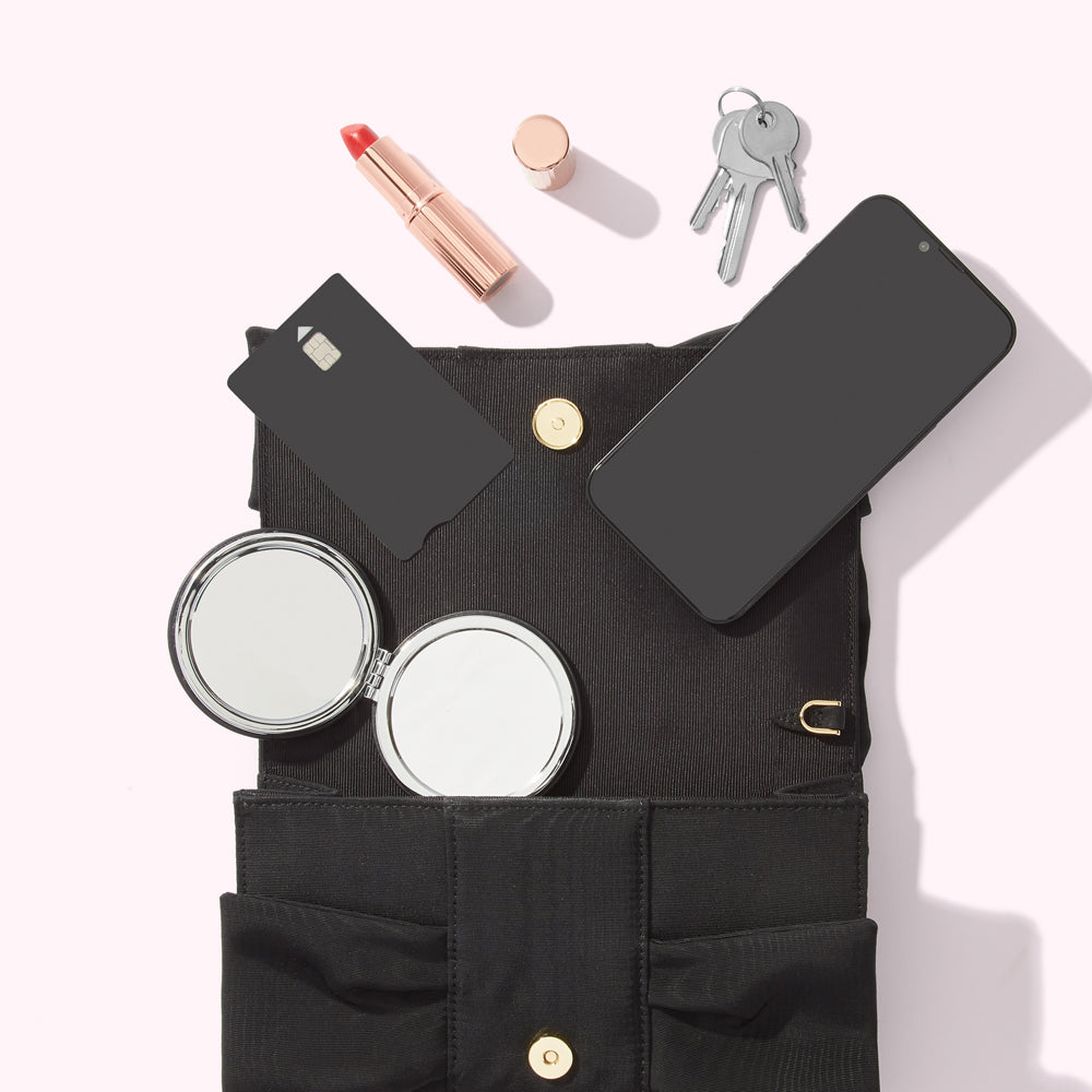 A black handbag open to reveal its contents: a smartphone, credit card, lipstick, compact mirror, and keys. The background is white.
