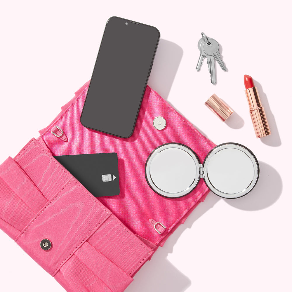 Pink clutch open to reveal a smartphone, credit card, compact mirror, lipstick, and keys.