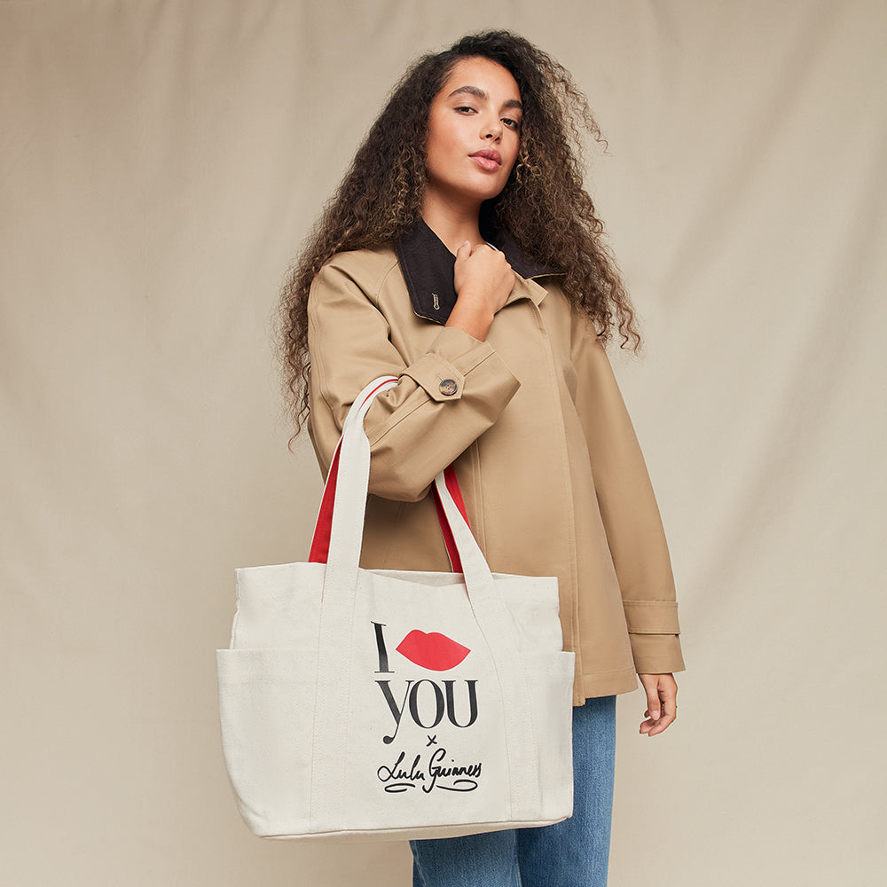 Woman holding a tote bag with 'I lip you x Lulu Guinness' text against a beige background