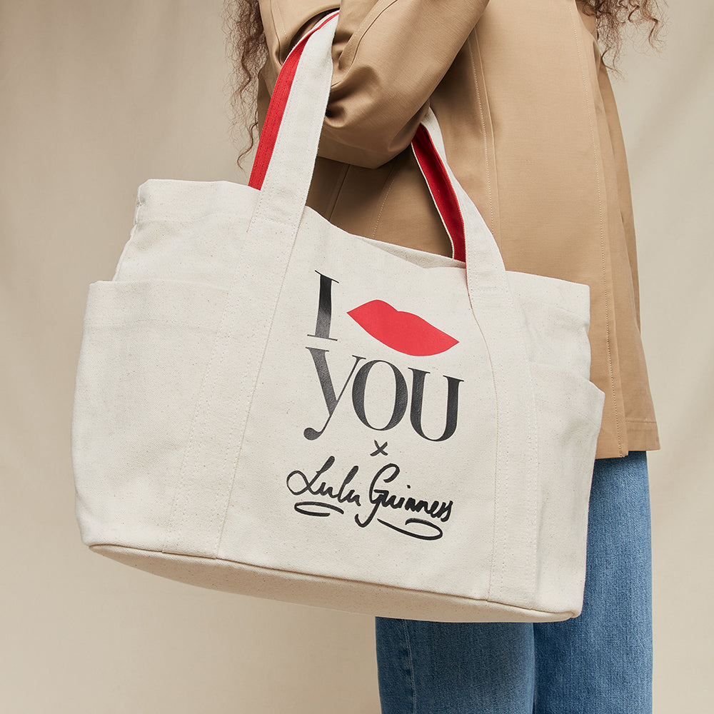 Tote bag with 'I lip you x Lulu Guinness' text and red lip print held by a person wearing a beige jacket.