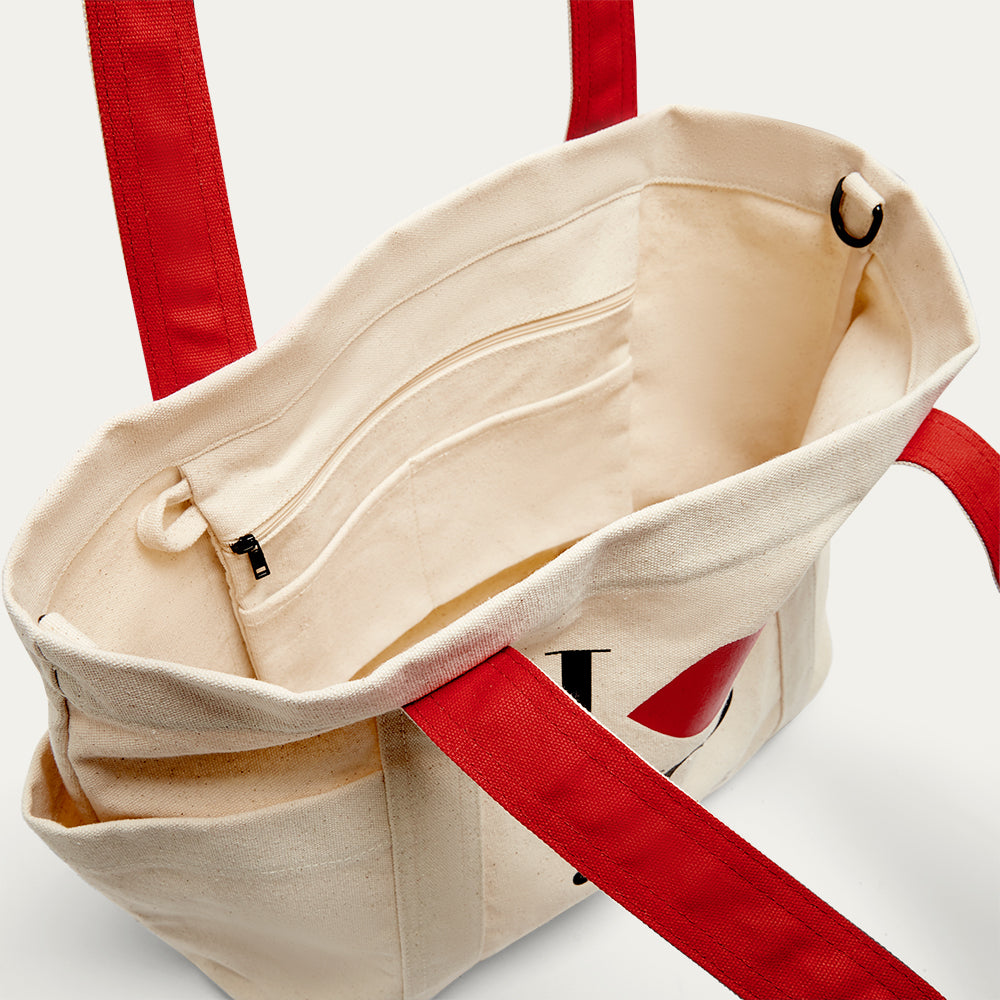 Beige tote bag with red straps on a light gray background open revealing a inside slip pocket, slip pocket and D-rings.