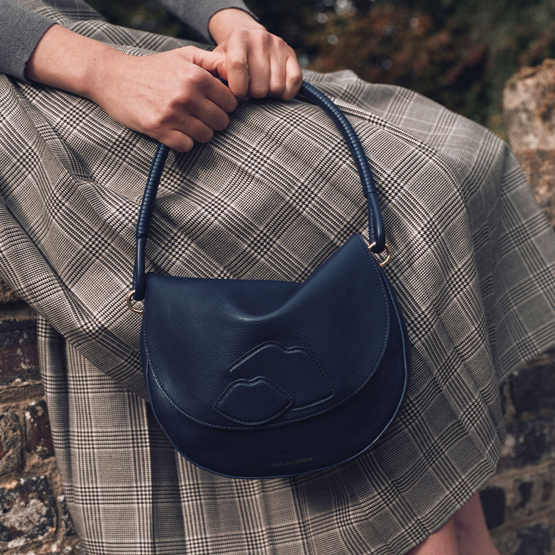 Person holding a navy blue handbag with a brand logo, wearing a plaid skirt.