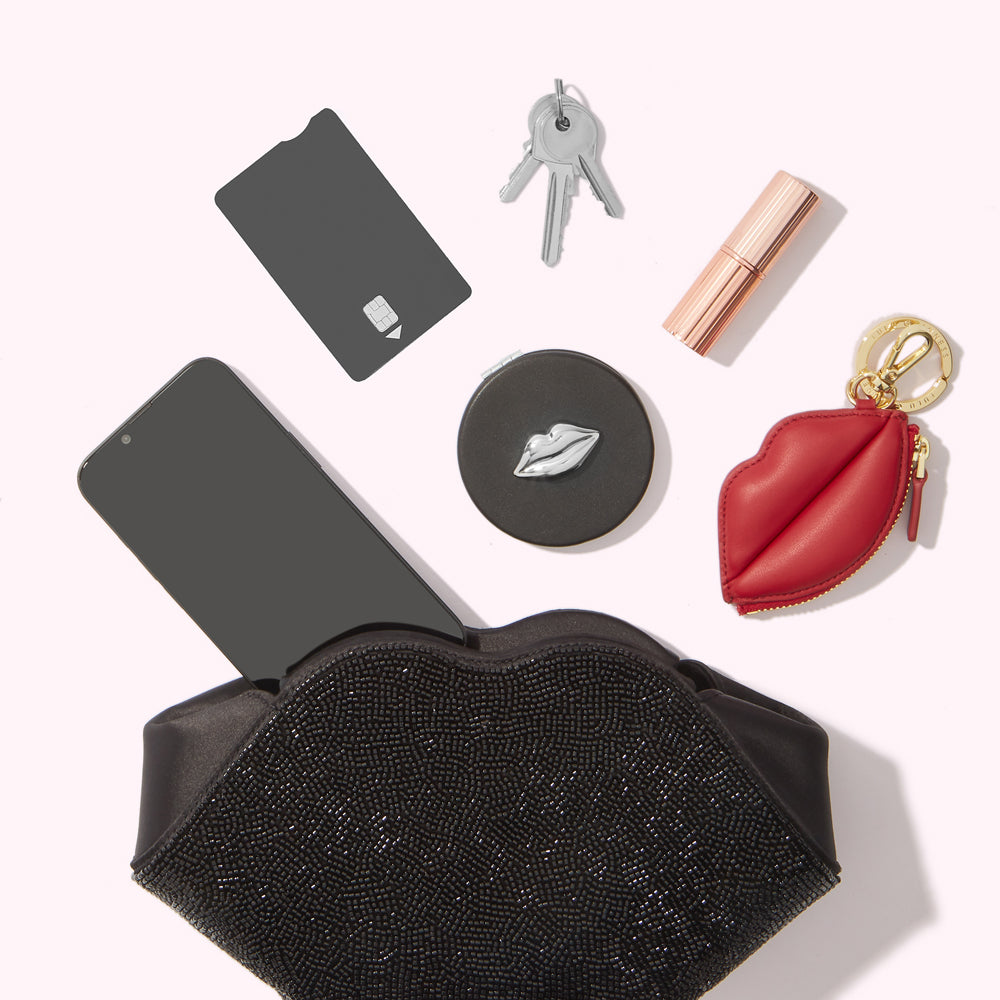 A black lip-shaped beaded clutch purse sits open with a smartphone, credit card, keys, lipstick, compact mirror, and red lip-shaped keychain inside, all on a white background.
