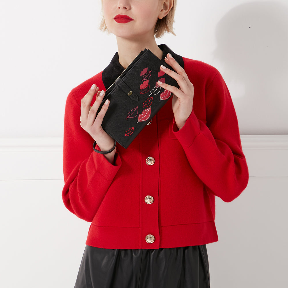 Woman in red jacket holding a black wallet with embroidered lips.
