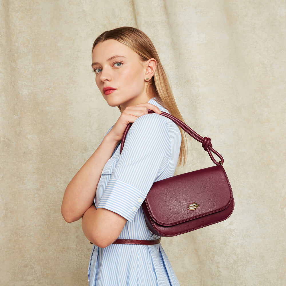 Woman holding a burgundy leather shoulder bag with gold lip details and a knotted handle against a beige background