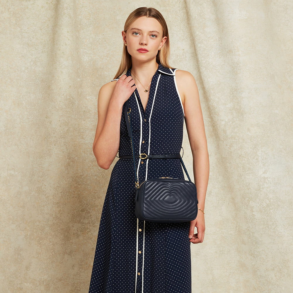 Woman in a navy polka dot dress wearing a navy leather quilted crossbody handbag against a beige background