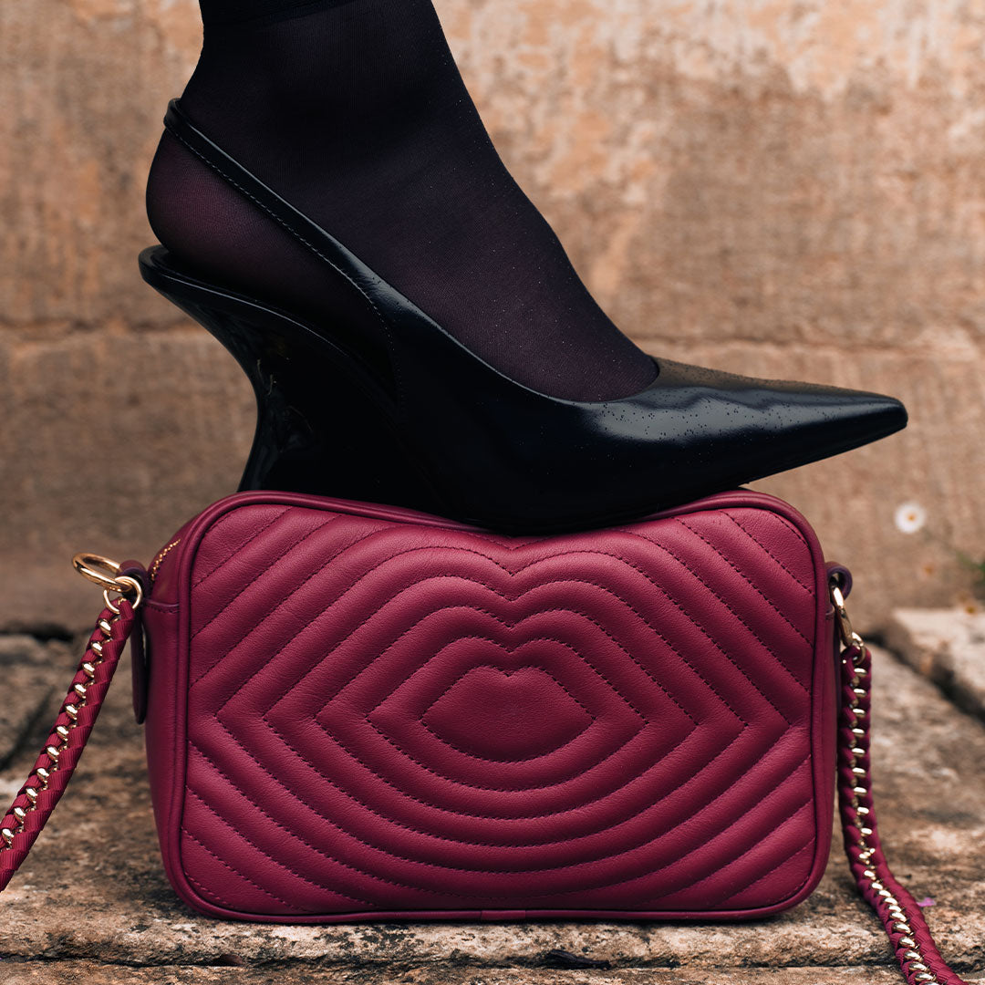 Burgundy quilted handbag with gold chain strap on stone surface, black high heel shoe on top.