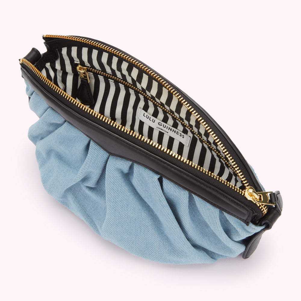 Light blue denim Lulu Guinness clutch purse open to reveal black and white striped interior and gold zipper.
