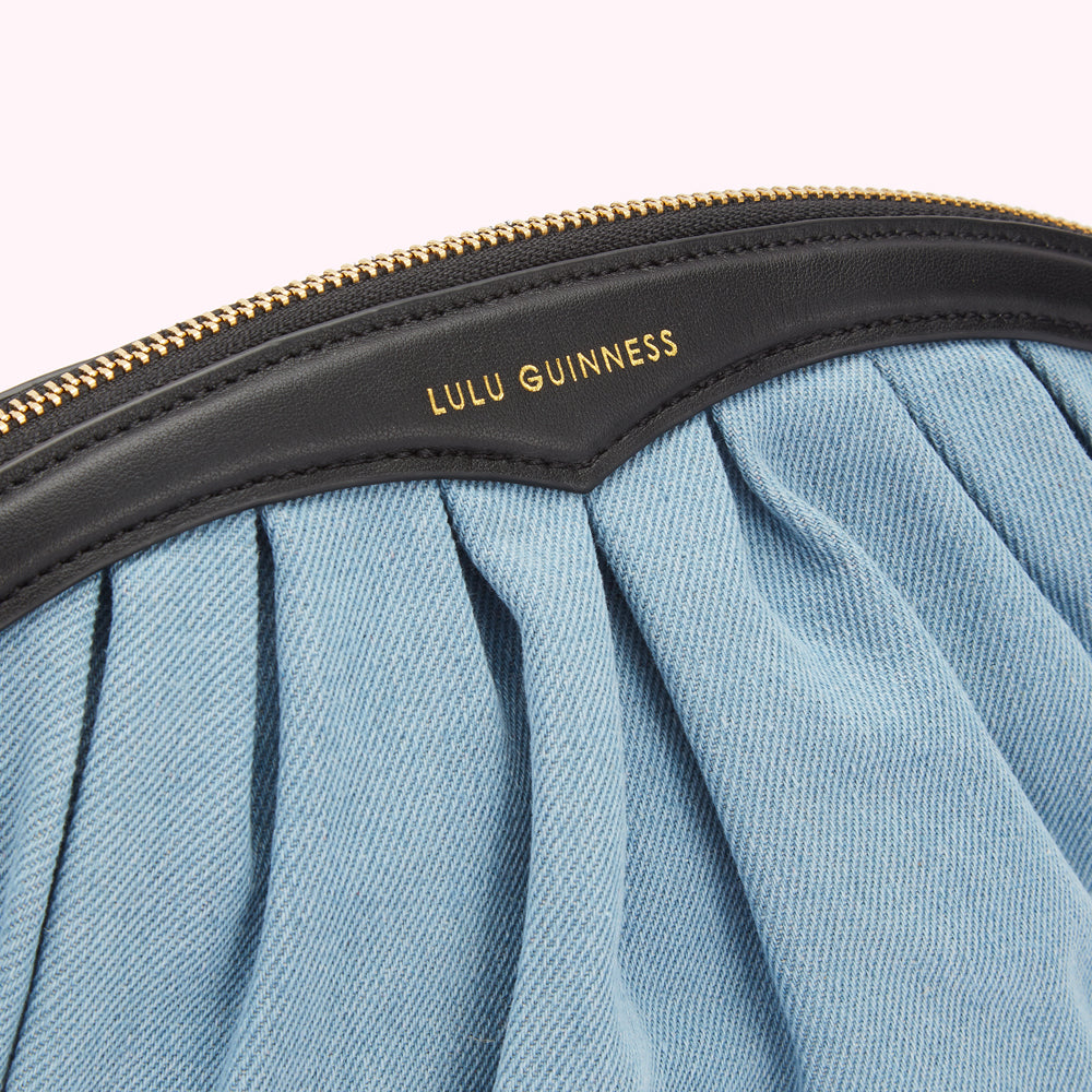 Close-up of a Lulu Guinness light blue denim clutch with a black leather top and gold zipper.
