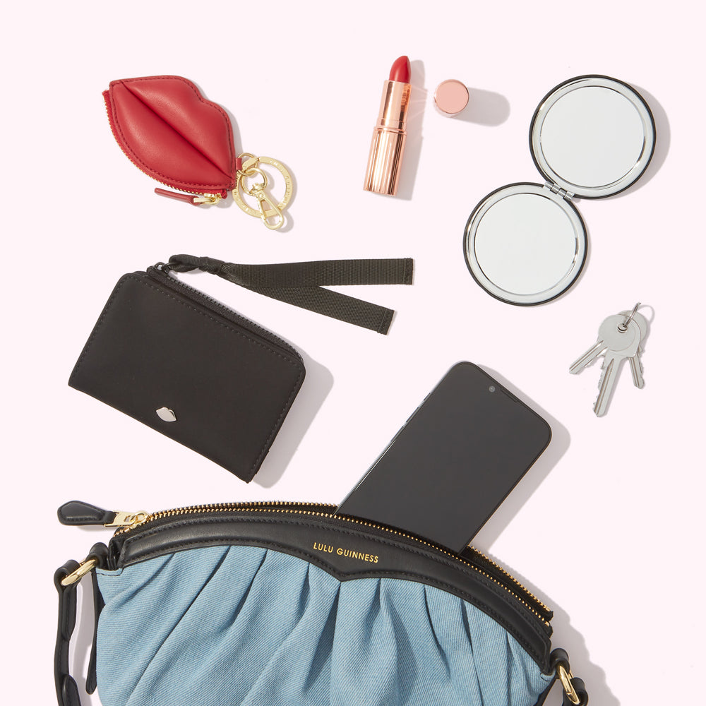 A light blue Lulu Guinness crossbody bag with a black card holder, a red lip-shaped keychain, a lipstick, a compact mirror, keys, and a smartphone inside on a white background.
