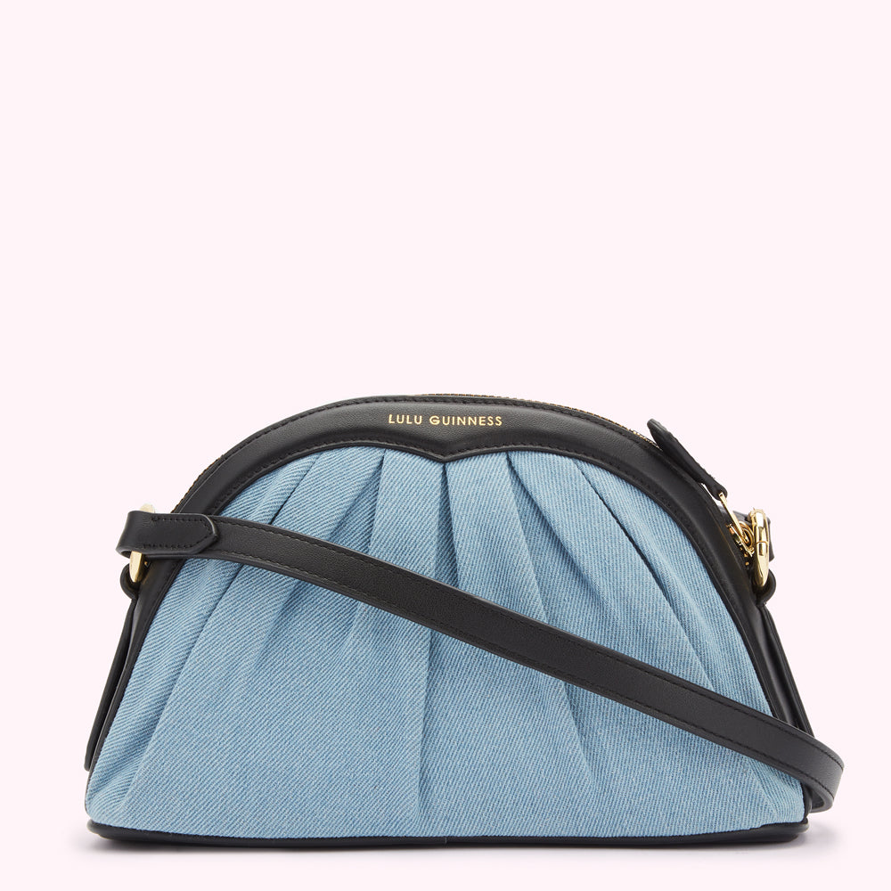 Light blue denim Lulu Guinness crossbody bag with black leather trim and gold hardware against a white background.
