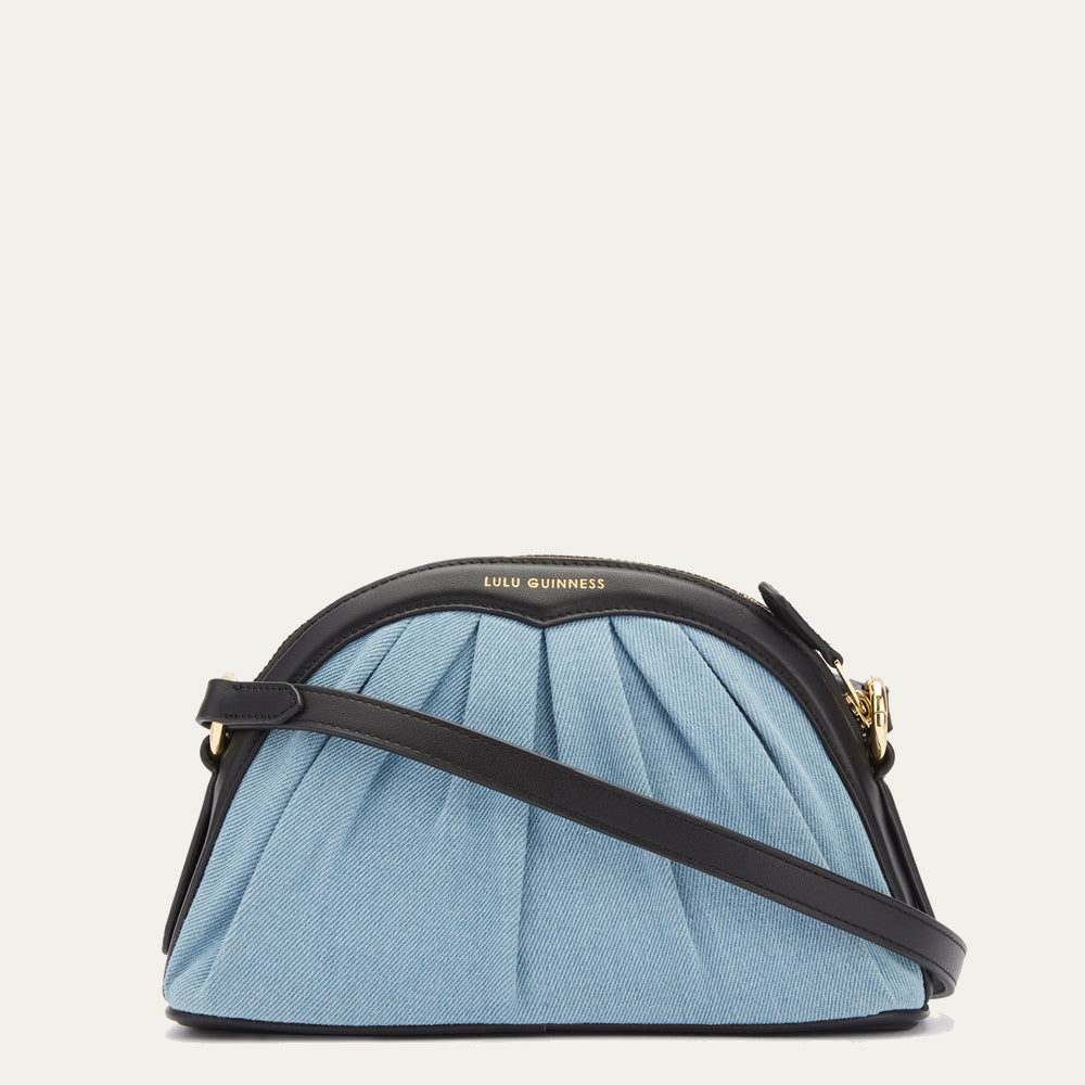 Light blue denim Lulu Guinness crossbody bag with black leather trim and gold hardware against a white background.