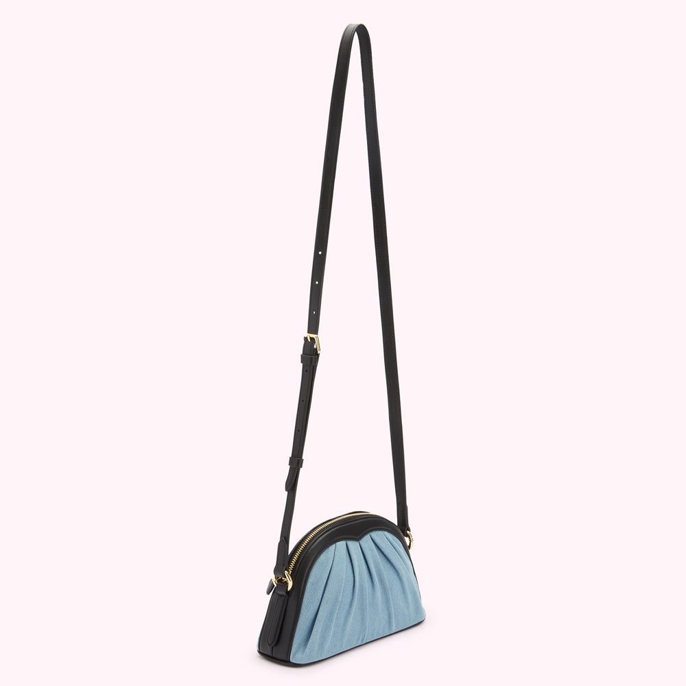 Half-moon shaped crossbody bag in light blue denim with black leather trim and long black strap against a white background.
