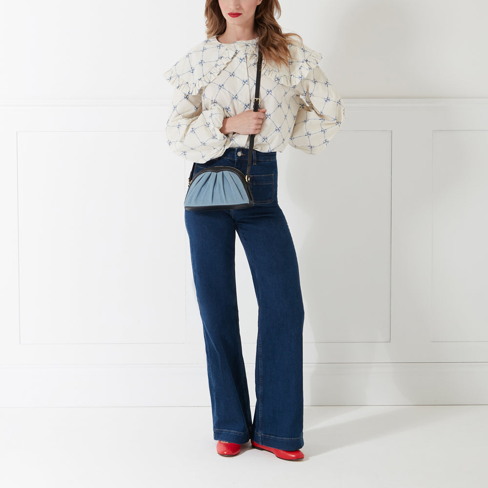 Woman wearing a light beige blouse, dark blue jeans, and red shoes, carrying a light blue and black crossbody bag against a white background.
