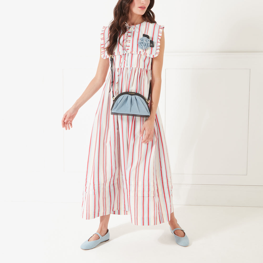 A model in a red striped dress and denim flat shoes wearing a denim and back crossbody bag and a denim rose shaped brooch. 
