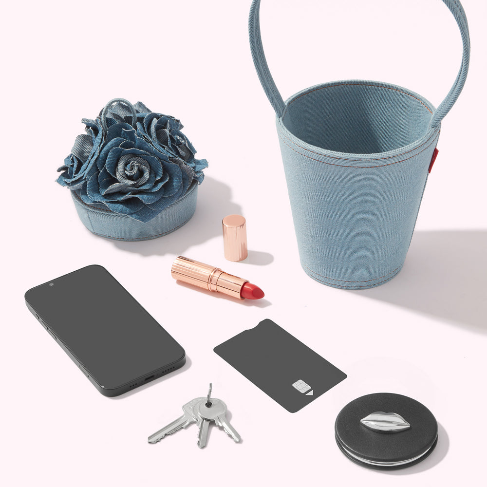 A flat lay of a denim bucket bag, a smartphone, keys, a credit card, lipstick, and a small container of denim roses on a white background.
