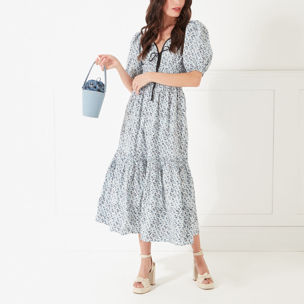 A full length image of a model with dark hair, red lipstick wearing a blue flower print dress holding a denim rose basket shaped bag in her hand.