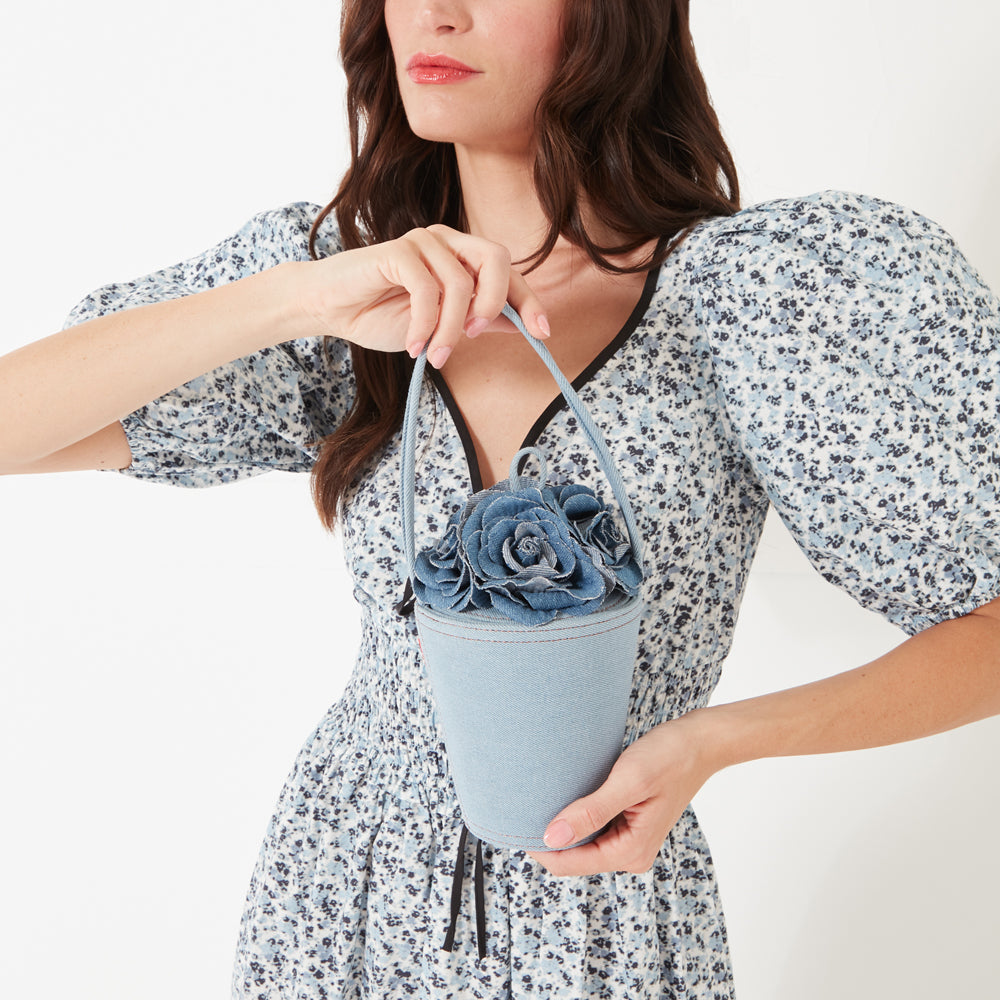 A close up image of a model with dark hair, red lipstick wearing a blue flower print dress holding a denim rose basket shaped bag.