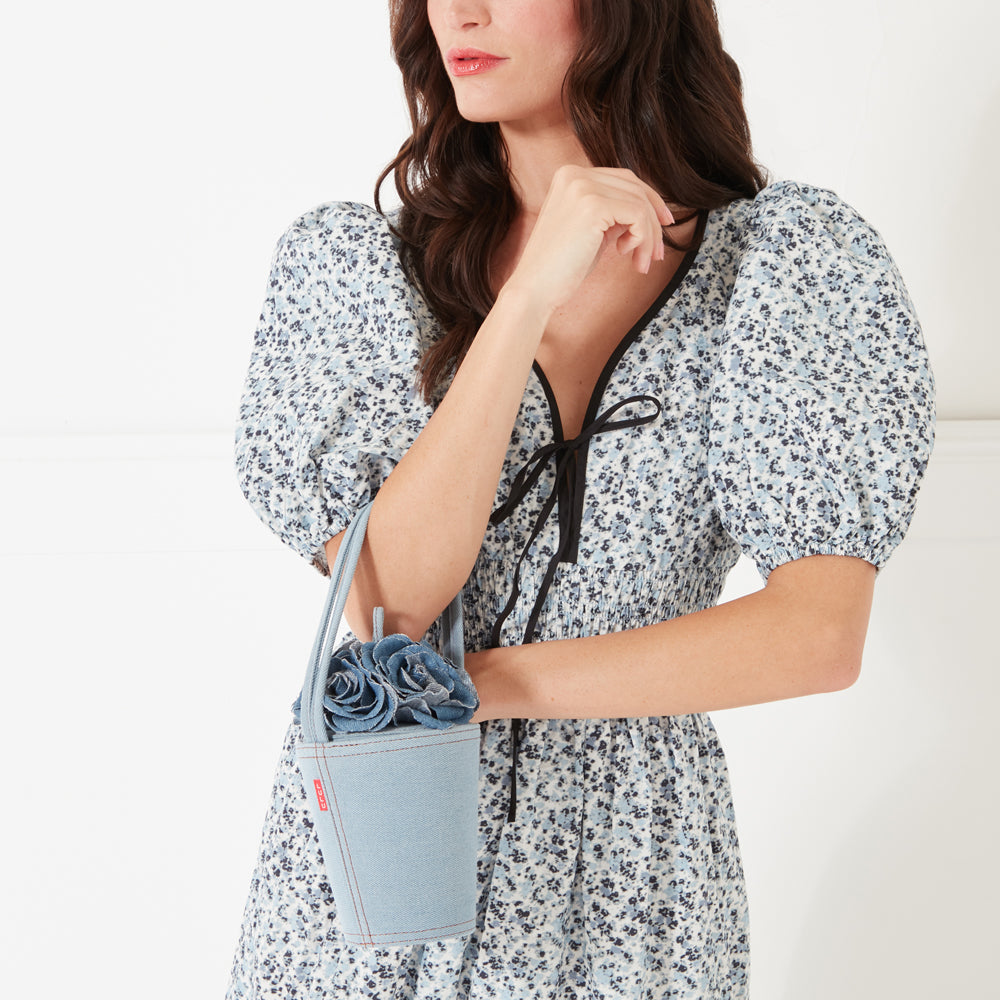 A close up image of a model with dark hair, red lipstick wearing a blue flower print dress holding a denim rose basket shaped bag on her arm.