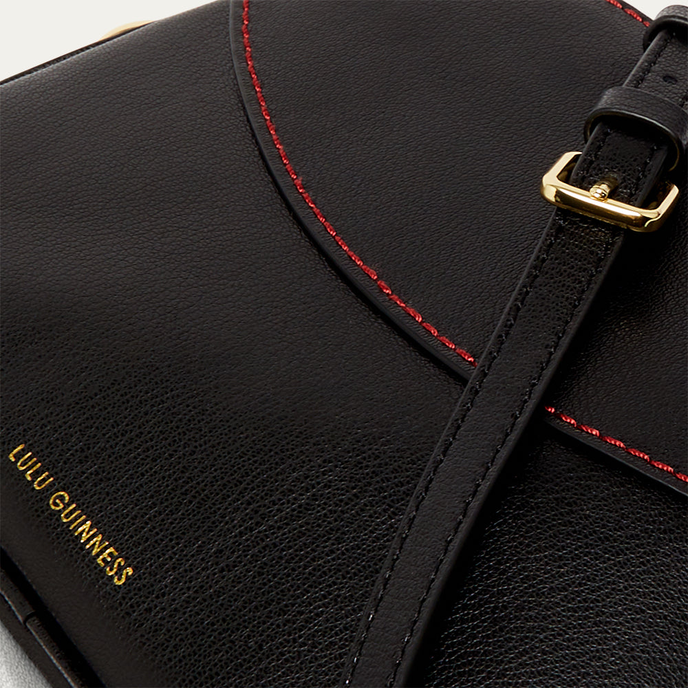 Details image of a black leather crossbody handbag with red stitching and gold buckle, featuring a printed 'Lulu Guinness' logo.
