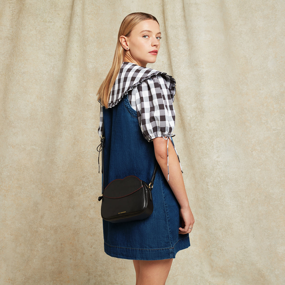 Woman wearing a denim dress, black and white gingham blouse and black leather crossbody bag against a beige background
