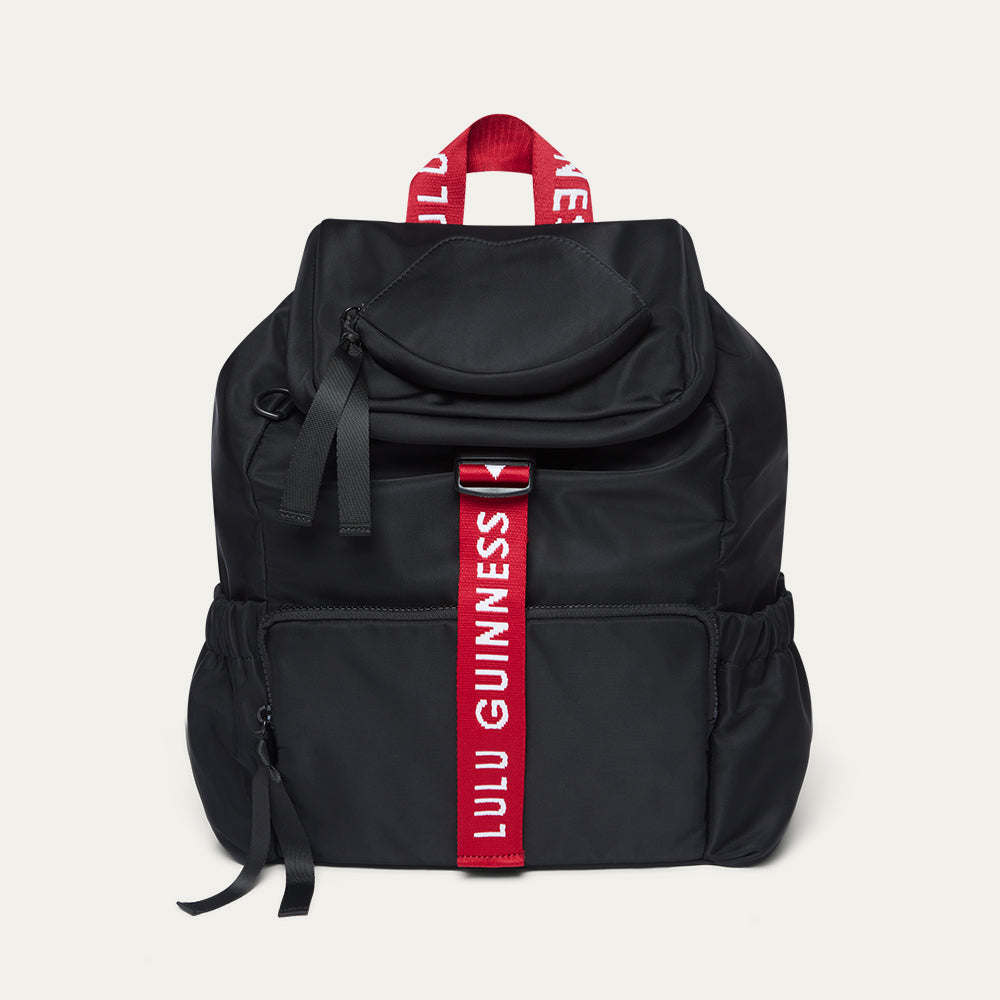 Black backpack with a red strap featuring 'Lulu Guinness' branding on a light grey background.