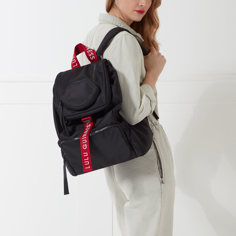 A woman in a light grey jumpsuit carries a black Lulu Guinness backpack with a red strap.
