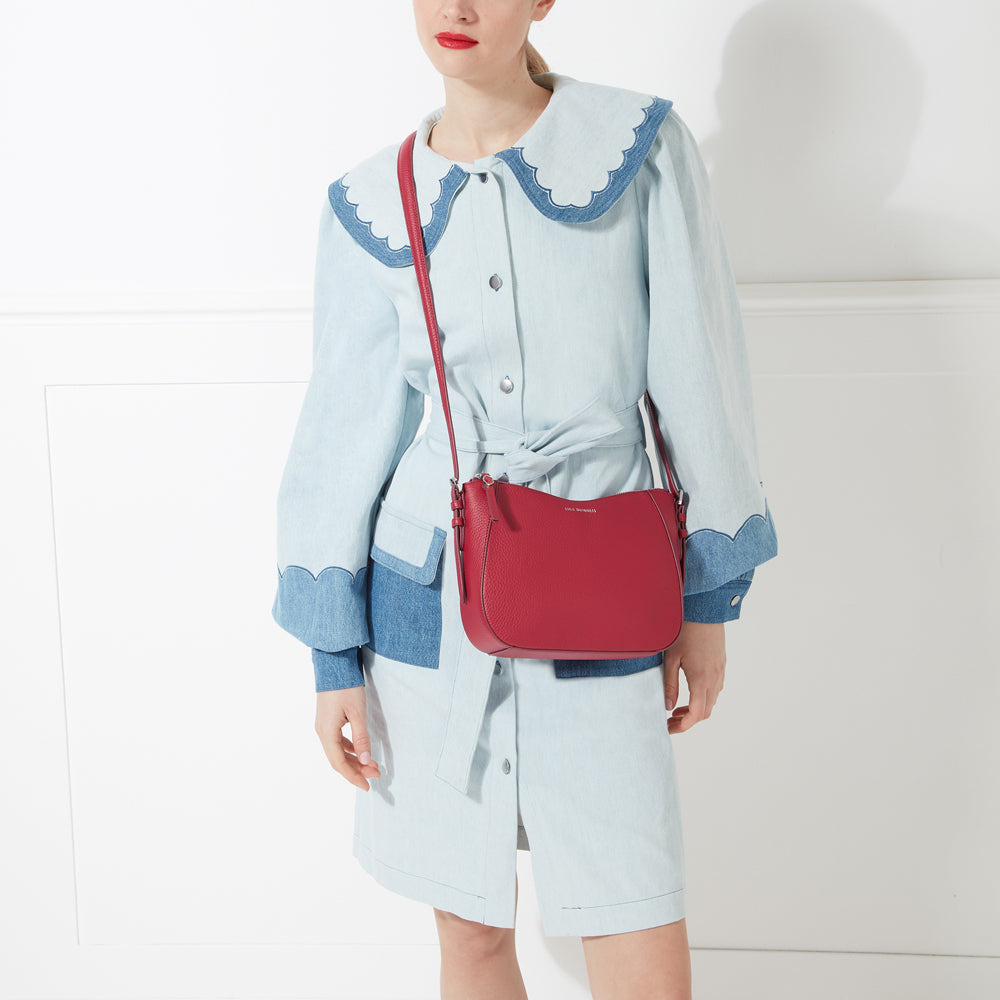 A red crossbody bag worn by a model in a light blue denim dress against a white background.
