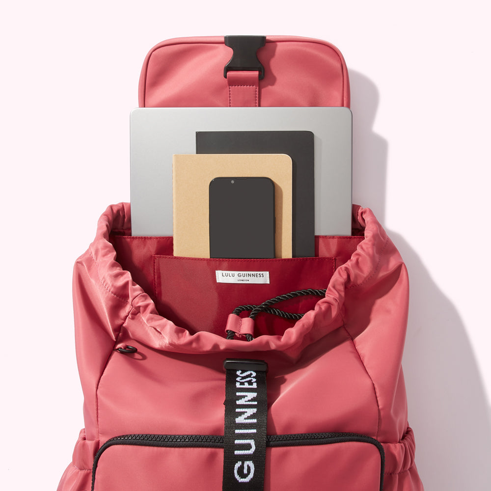 A dusty rose Lulu Guinness London backpack open to reveal a laptop, notebook, phone, and other stationery items inside.
