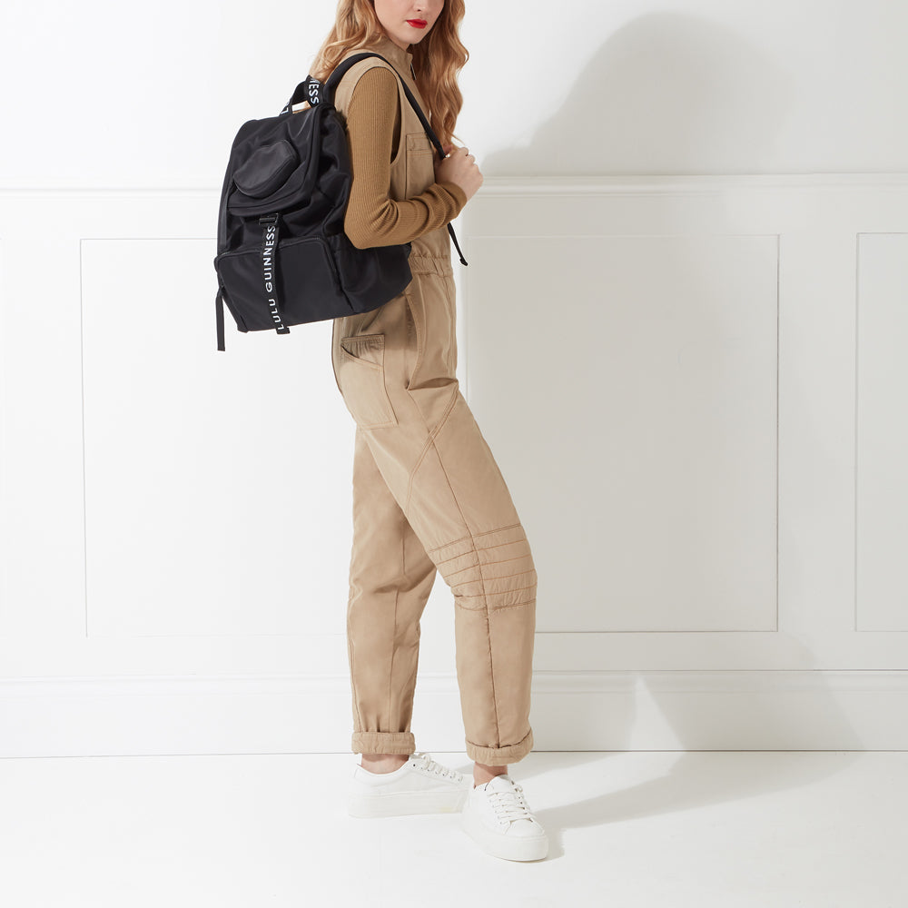 Woman wearing beige jumpsuit and white sneakers, carrying a black Lulu Guinness backpack.
