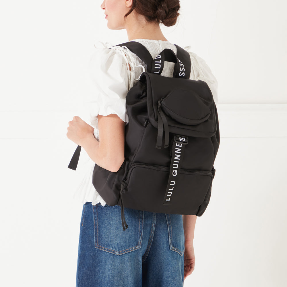 Back shot of a model wearing a white top and denim jeans wearing a black nylon backpack with 'Lulu Guinness' branded straps on her back.
