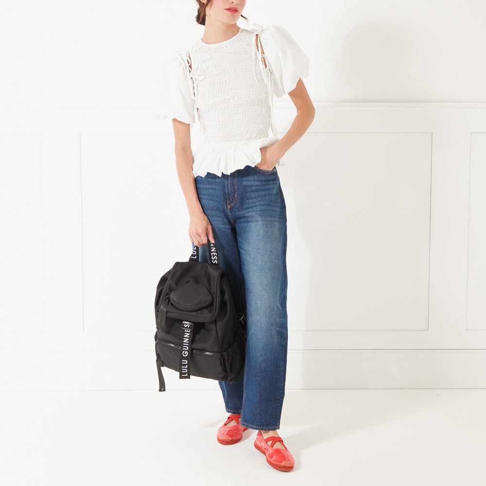 Full length image of a model wearing jeans, a white top and red shoes holding a black nylon backpack with 'Lulu Guinness' branded straps down in her hand.