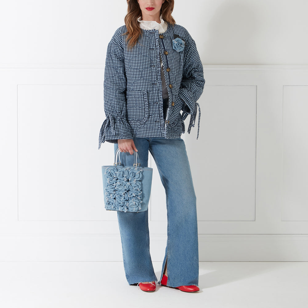 Woman in blue gingham jacket, light wash jeans, and red shoes carries a light blue denim tote bag adorned with fabric roses. The background is white.
