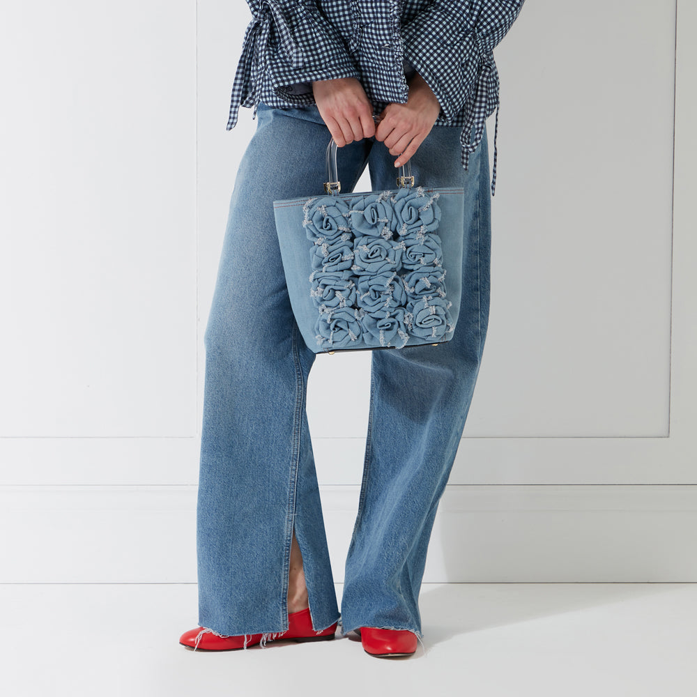 A woman in blue jeans and a gingham shirt holds a light blue denim handbag adorned with fabric roses. She is wearing red flats. The background is white.
