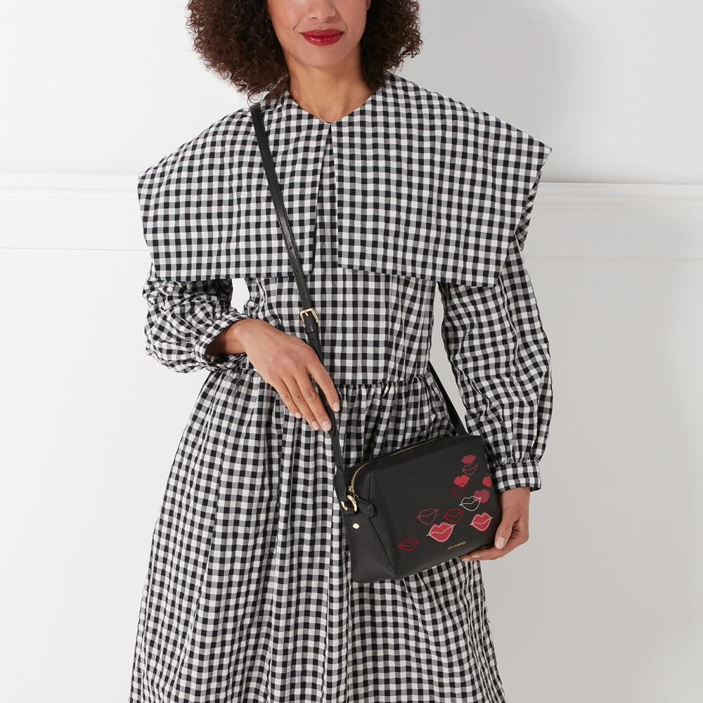 A woman in a black and white gingham dress holds a black crossbody bag with red lips printed on it.