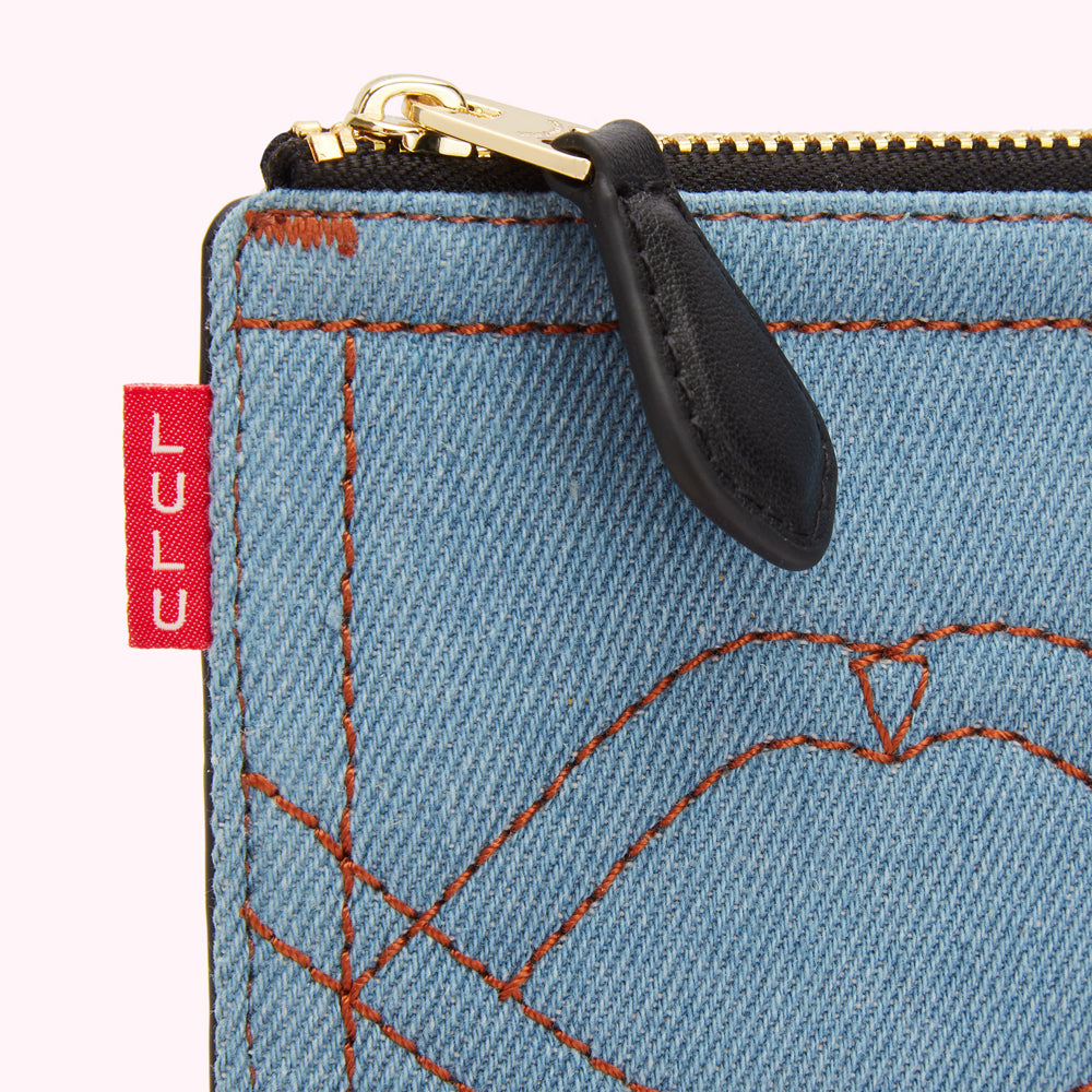 Close-up of a light blue denim zipper pouch with orange stitching and a black leather zipper pull. A red label is partially visible.
