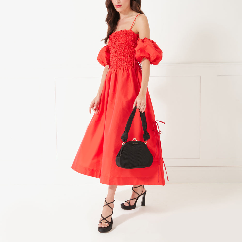 A full length image of a model with dark hair and red lipstick wearing a red occasion dress and black heeled shoes holding a black satin plisse clutch bag by its handle.