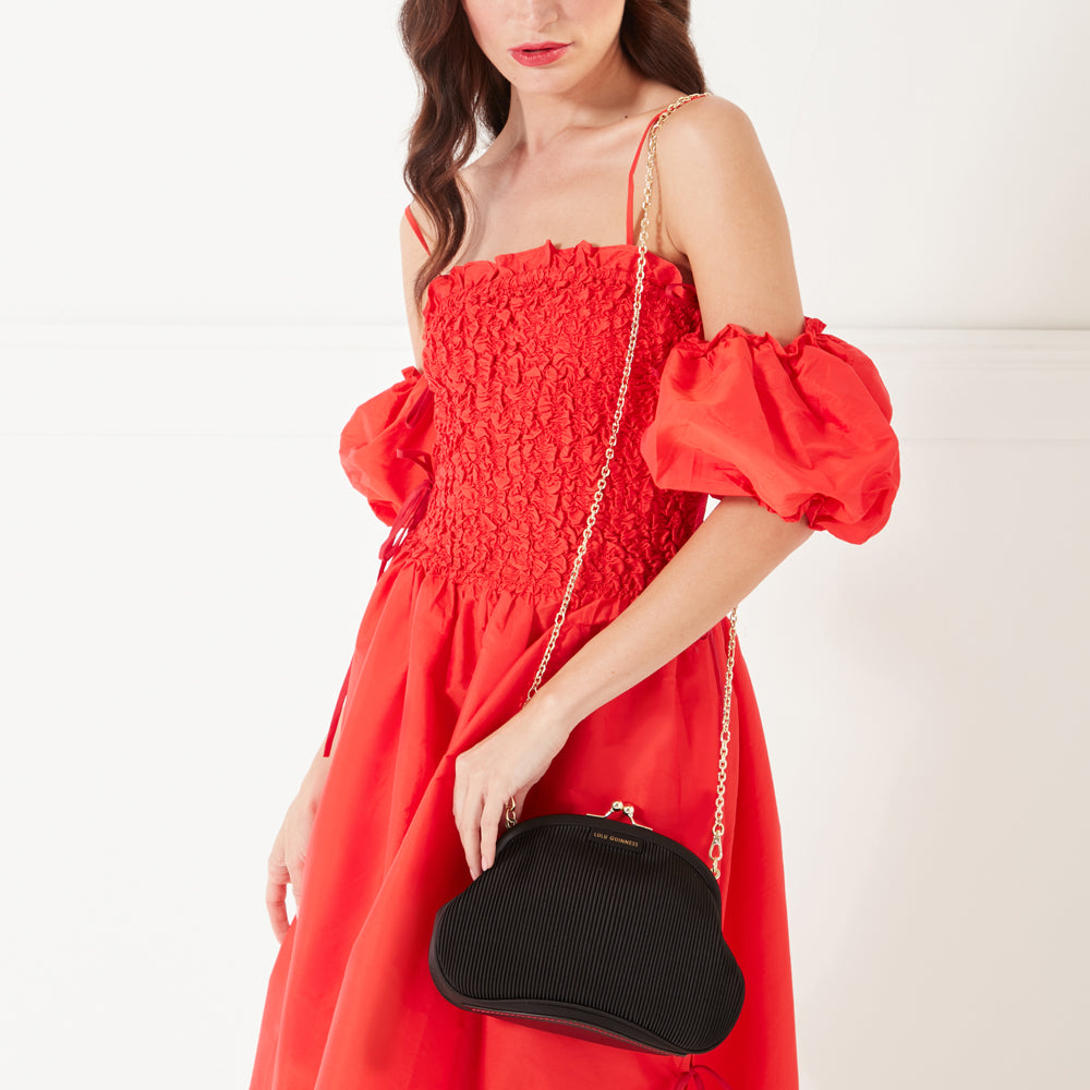 A model with dark hair and red lipstick wearing a red occasion dress and a black satin plisse clutch on her shoulder.