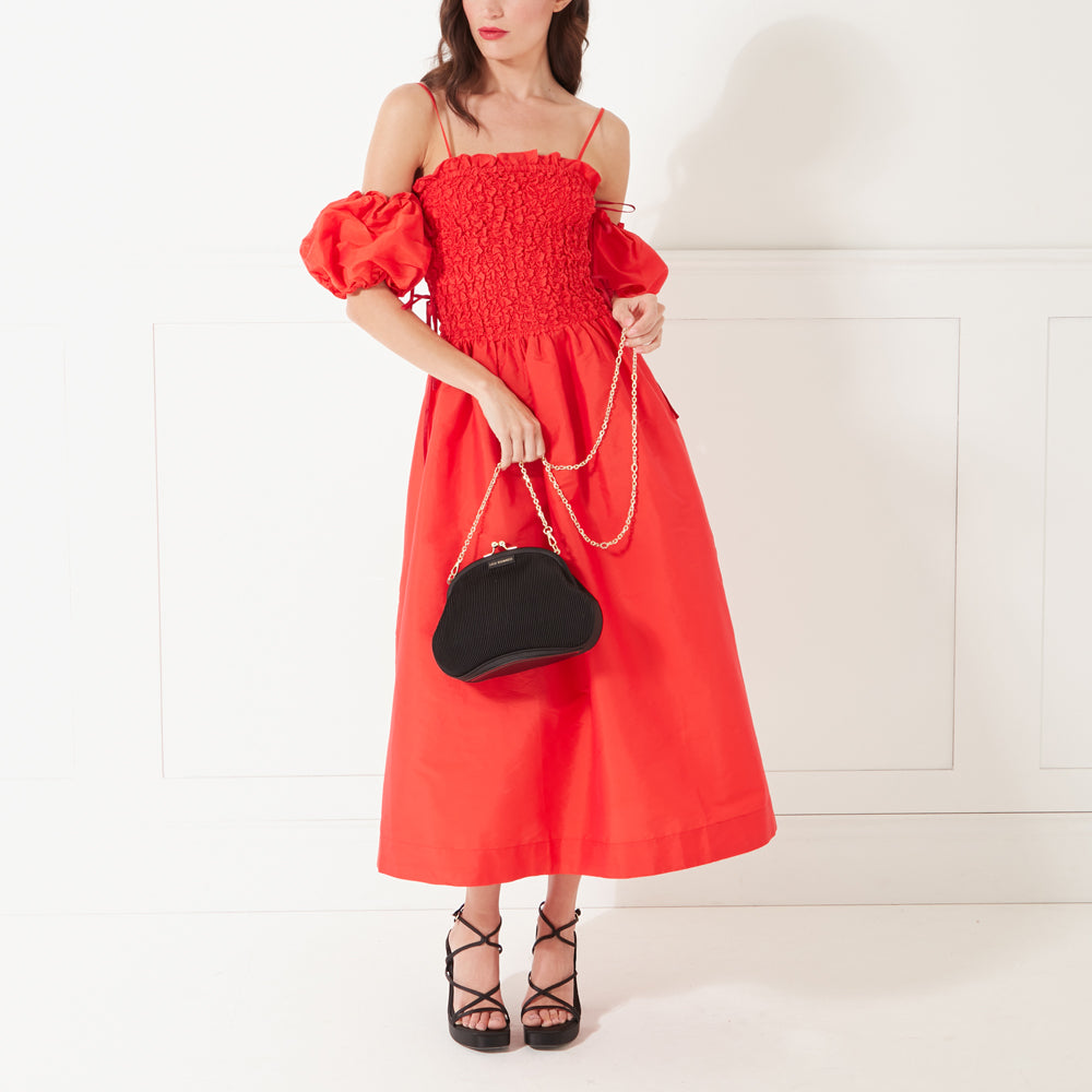 A full length image of a model in a red occasion dress and black heels holding a black satin plisse clutch bag by its gold chain.