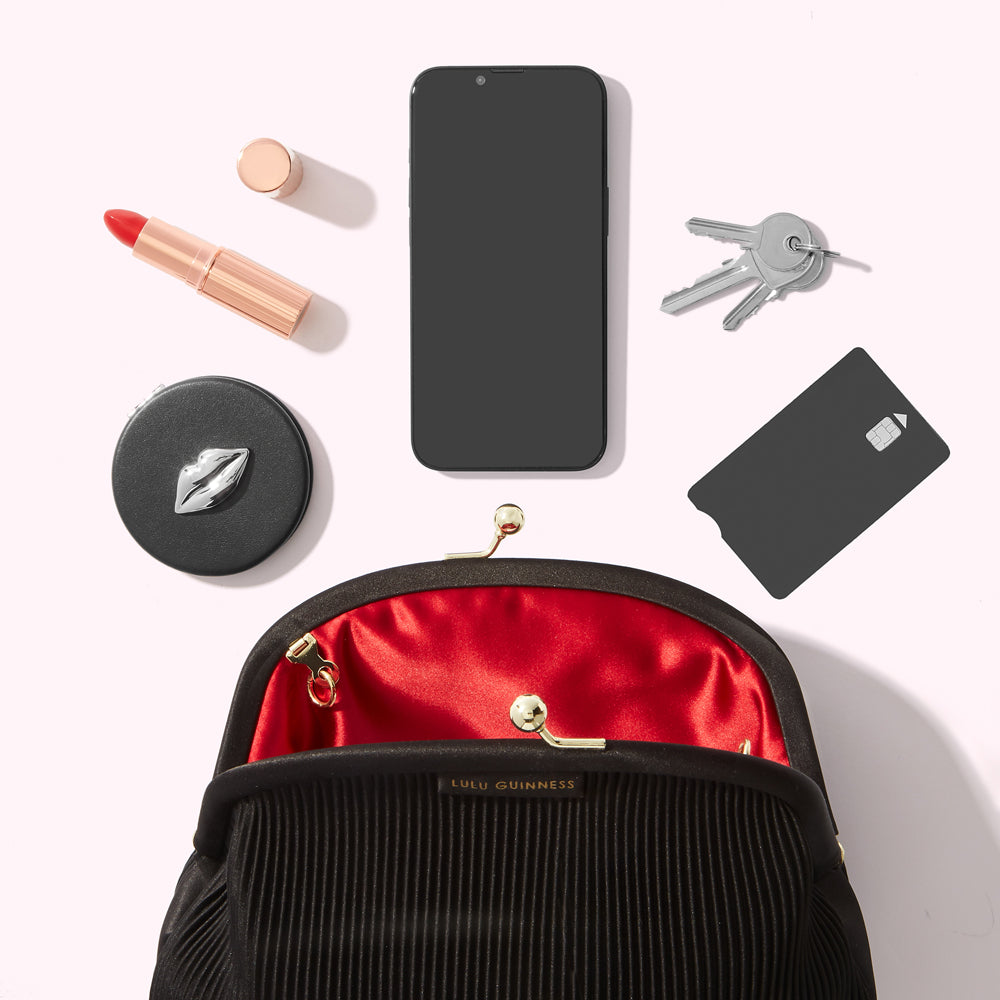 Flatlay of a black Lulu Guinness purse with red satin interior, a smartphone, keys, lipstick, compact mirror and credit card on a white background.
