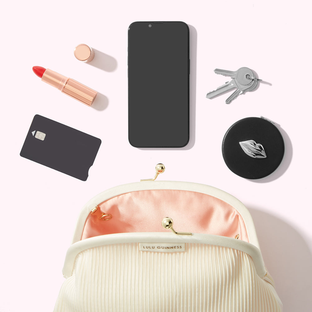 A cream-colored Lulu Guinness purse lies open, revealing a peach-coloured interior. Surrounding it are a smartphone, keys, a compact mirror with a lip design, a credit card, and lipstick. The background is white.
