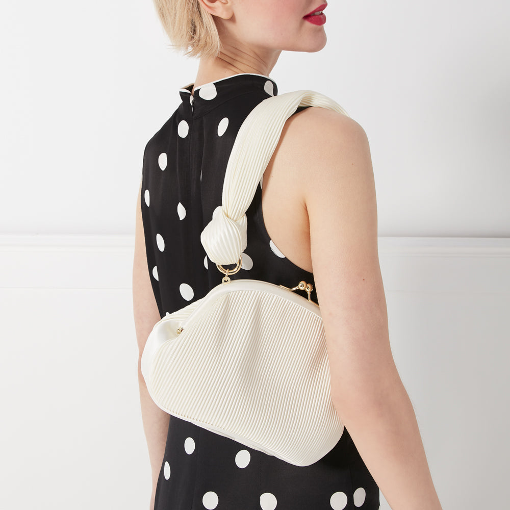 A woman in a black polka dot dress carries a pleated off-white handbag with a knotted strap.
