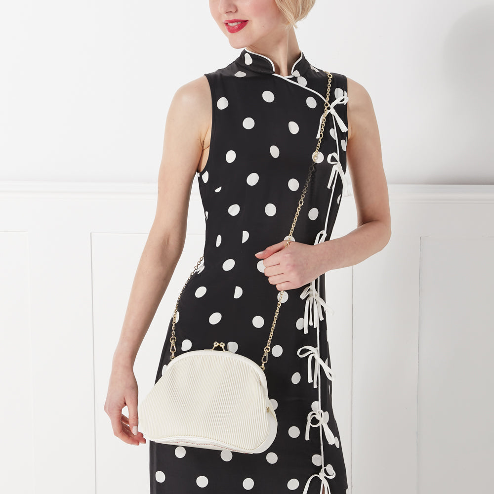 Woman in black and white polka dot dress holding a white purse with gold chain strap.
