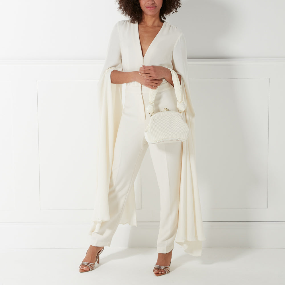 Woman in a white jumpsuit and cape sleeves holds a white pleated handbag against a white background.
