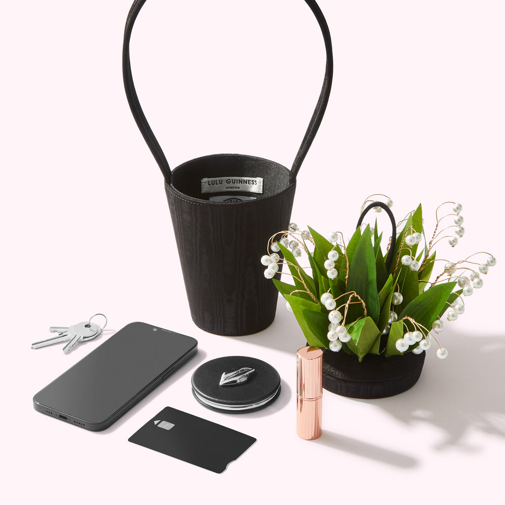 A black Lulu Guinness handbag, a smartphone, keys, a credit card, a compact mirror, and a lipstick are arranged on a white surface next to a lily of the valley arrangement in a small black bag.