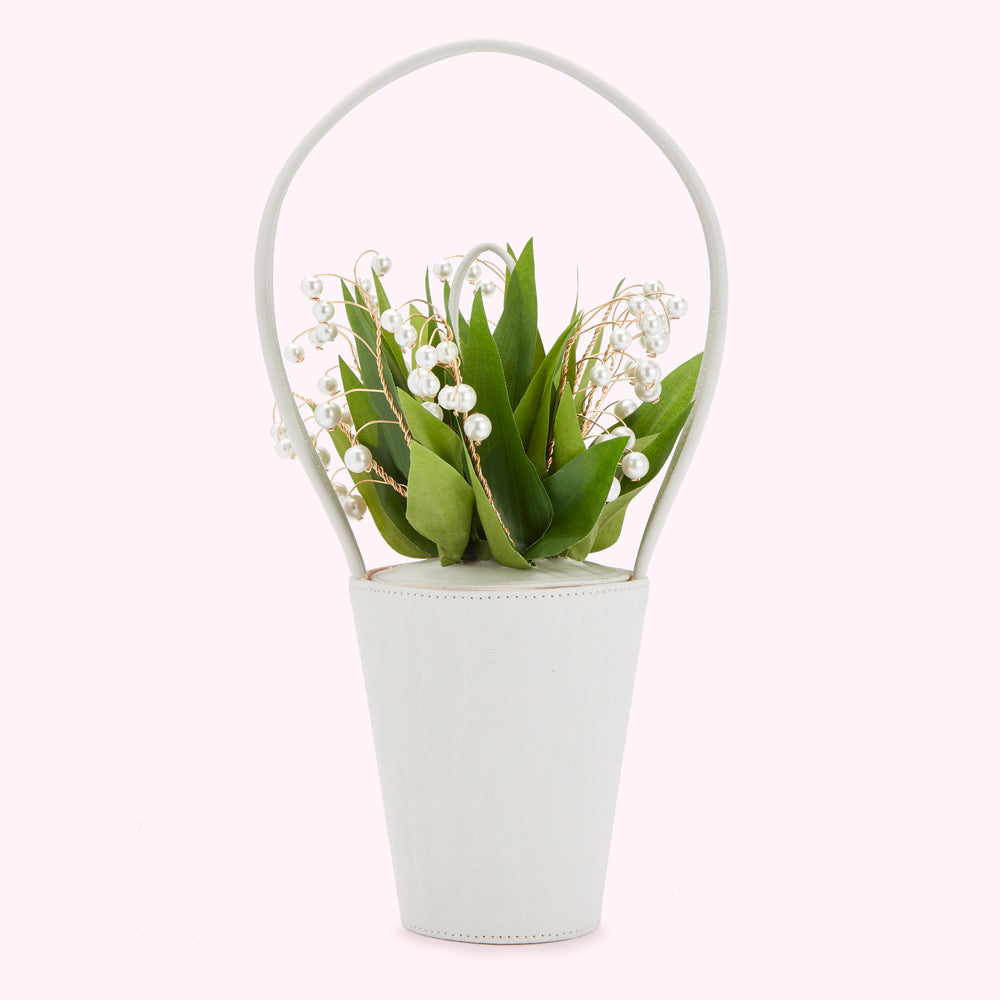 A ivory white, bucket-shaped handbag with a handle sits on a white background. A bouquet of lily of the valley accented with pearls is nestled inside the bag.
